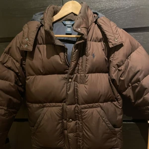 Brun dunjacka från Polo Ralph Lauren - Säljer en brun dunjacka från Polo Ralph Lauren i storlek L i barn storlek men passar mig som är 175 och har s/ m. Jackan är quiltad och har en dragkedja samt knappar framtill. Perfekt för kalla vinterdagar med sin varma och sköna passform. Den är i bra skick med några mindre tecken på användning. Passar både till vardags och lite finare tillfällen.