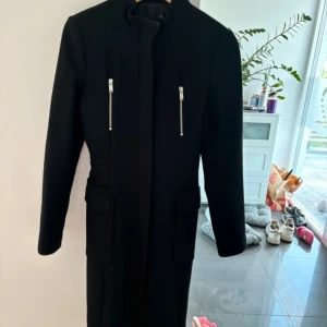 Svart kappa med dragkedjor - Säljer en stilren svart kappa i mycket bra skick. Den har långa ärmar och två snygga dragkedjor framtill. Perfekt för höst och vinter, och ger en elegant look. Passar både till vardags och festliga tillfällen. Super fint skick! 