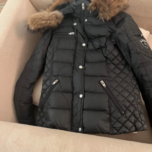 Svart quiltad dunjacka med pälsdetaljer - Säljer en snygg svart dunjacka med quiltat mönster och pälsdetaljer på huvan. Jackan har dragkedja och knappar framtill samt praktiska fickor med dragkedjor. Perfekt för kalla vinterdagar! 🖤rock and blu med äkta päls