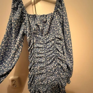 Blå blommig klänning - Säljer en superfin blå klänning med små vita blommor. Den har långa ärmar och en snygg rynkad design framtill som ger en cool look. Perfekt för vår och sommar! 🌸