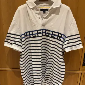 Säljer en snygg vit pikétröja från Tommy Hilfiger med marinblå ränder och texten 'HILFIGER' över bröstet. Tröjan har en klassisk krage och korta ärmar, perfekt för en avslappnad stil. Den är i mycket bra skick och passar både till vardags och festliga tillfällen.