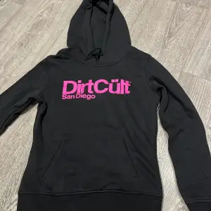 Säljer en svart hoodie från DirtCült med rosa tryck 'San Diego' på framsidan. Den har en klassisk känguruficka och justerbar huva. Perfekt för en avslappnad stil eller kyliga dagar. Skön och stilren design!