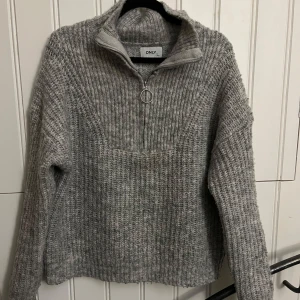 Grå stickad tröja från ONLY - Mysig grå stickad tröja från ONLY med dragkedja i halsen. Perfekt för kyliga dagar med sin ribbade design och långa ärmar. Passar både till vardags och mysiga hemmakvällar. 🧶