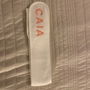 Vitt diadem/ hårband från CAIA - Säljer ett vitt diadem/hårband från CAIA i mjukt material. Perfekt för att hålla håret borta vid sminkning eller ansiktsvård. Diademet/hårbandet har en enkel design med broderad logga i rosa. Använd det för en bekväm och stilren look!