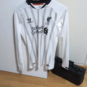 Säljer en vit långärmad Liverpool-tröja från Warrior i mycket bra skick. Tröjan har svarta ränder och LFC-loggan broderad på bröstet. Perfekt för alla Liverpool-fans som vill ha något unikt i garderoben. Passar bra till både match och vardag!