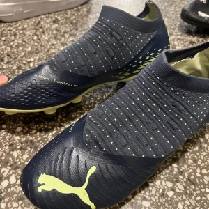 Puma fotbollsskor - Säljer ett par grymma Puma fotbollsskor i mörkblått med neongula detaljer. Skorna har en strumpliknande passform och är perfekta för spel på gräs. De har en snygg design med små prickar och det klassiska Puma-loggan på sidan. Sulan är utrustad med dobbar för optimalt grepp. Perfekta för dig som vill dominera på planen!