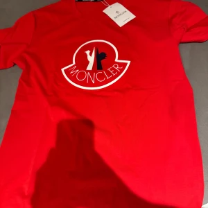 Röd t-shirt från Moncler - Säljer en snygg röd t-shirt från Moncler i bomull. Den har en stor logga på bröstet i vitt och blått. Perfekt för en casual look eller att styla upp med en jacka. T-shirten är kortärmad och i nytt skick. Passar perfekt till sommaren eller våren!