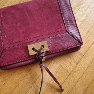 Vinröd axelväska med mockadetaljer - Snygg vinröd axelväska i skinn med mockadetaljer och ormskinnsmönster. Väskan har en elegant knytning framtill och en kedjedetalj på axelremmen. Perfekt för både vardag och fest!