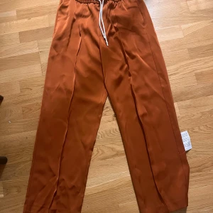 Bruna byxor med snörning - Säljer ett par snygga orange byxor i mycket bra skick. De har en loose passform och är högmidjade med snörning i midjan för extra komfort. Perfekta för höst och vår, och de har en stilren look som funkar både till vardags och fest.