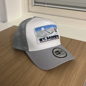 Vit och grå keps från New Era - Snygg vit och grå keps från New Era med ett coolt St. Moritz-märke framtill. Perfekt för en avslappnad stil eller för att skydda mot solen. Kepsen har en justerbar baksida för perfekt passform och ett nätmaterial som andas. Följer även med en caphunters påse vid köp, kepsen är aldrig använd mer än att jag testat den och eftersom den inte var min stil så säljer jag den istället. Därmed är klistmärken fortfarande på, om priset inte passa lägg gärna ett bud👍