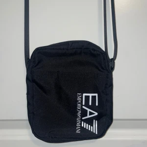 Svart axelväska från Emporio Armani - Snygg svart axelväska från Emporio Armani med justerbar rem. Perfekt för vardagsbruk eller en kväll ute. Väskan har en stilren design med logotypen tryckt på framsidan. Den är lätt och smidig att bära med sig.