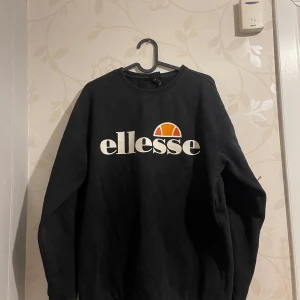 Svart sweatshirt från Ellesse - Säljer en svart sweatshirt från Ellesse i bra skick. Den har en stor logga på framsidan i vitt och orange. Perfekt för höst och vinter, med långa ärmar och en bekväm passform. Passar både till vardags och chillkvällar. Passar lika bra på storleken S som M.