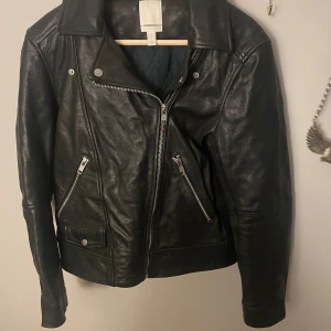Svart skinnjacka från H&M - Snygg svart skinnjacka från H&M med dragkedja och knappar.