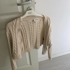 Beige stickad kofta från Monki - Säljer en snygg beige stickad kofta från Monki. Koftan har ett flätat mönster och är kort i modellen. Perfekt för lager-på-lager-stil och passar till många olika outfits.