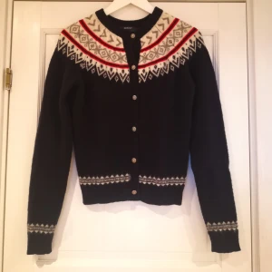 Mörkblå lammulskofta från Gant - Säljer en mörkblå kofta från Gant i lammull med ett vackert mönster i rött, vitt och grått runt halsen. Koftan har långa ärmar och knappar i metall framtill. Perfekt för kyliga dagar!