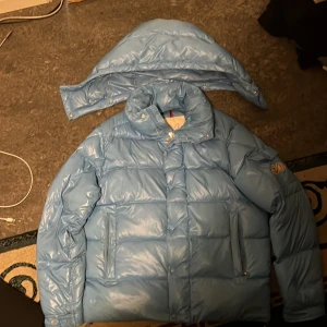 Ljusblå maya monclear jacka - Säljer en ljusblå dunjacka från Moncler med avtagbar huva och dragkedja. Jackan har långa ärmar och Moncler-logga på ärmen. Perfekt för kyliga dagar.