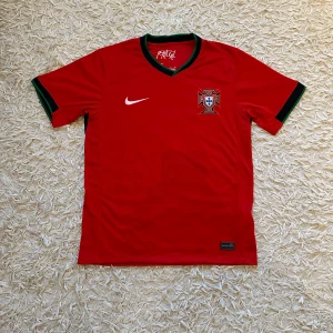 Portugal fotbollströja-  med originella lappar  - Säljer en röd fotbollströja från Nike med Portugals emblem på bröstet. Tröjan har korta ärmar och gröna detaljer vid ärmslut och krage. Helt ny, oanvänd med alla originella lapparna kvar, perfekt för sommaren och för dig som gillar fotboll! 🇵🇹