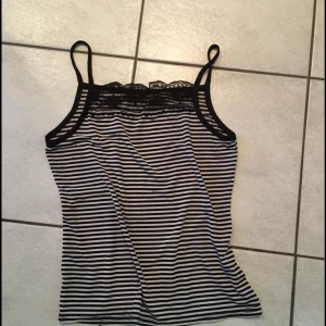 Randig linne med spetsdetaljer - Snyggt randigt linne i svart och vitt med spetsdetaljer vid halsringningen. Linnet har smala axelband och en bekväm passform, perfekt för varma dagar.