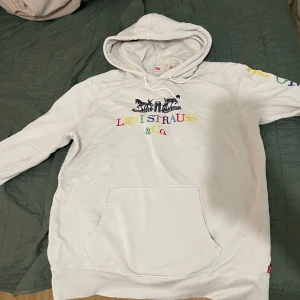 Vit hoodie från Levi's - Säljer en vit hoodie från Levi's med färgglad logotyp på bröstet. Den har en klassisk känguruficka och justerbar huva med dragsko. Perfekt för en avslappnad stil.