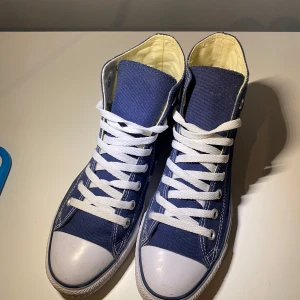 Blå höga sneakers från Converse - Snygga blå höga sneakers från Converse med klassisk vit gummisula och snörning. Perfekta för en avslappnad stil. De har det ikoniska Converse-märket på sidan. Pris kan diskuteras