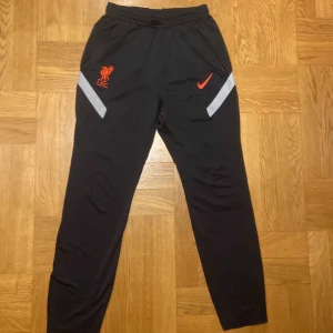 Liverpool x Nike träningsbyxor - Säljer ett par svarta Nike Dri-FIT träningsbyxor med Liverpool FC-logga. Byxorna har elastisk midja och är dekorerade med röd Nike och Liverpool-logga och vita detaljer. Perfekta för fotbollsträning eller annan sportaktivitet. Ett litet hål i fickan annars 👍finns lite slitningar här o där dock se bild. 