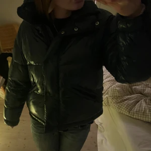 Mörkgrön glansig pufferjacka - Säljer en mörkgrön glansig pufferjacka med  dragkedja. Jackan har en glansig yta och är perfekt för kyligare dagar. Den har långa ärmar och en bekväm passform. Den är varm och gosig till vintern 