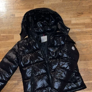 Svart jacka  från Moncler - Snygg svart jacka från Moncler med glansig finish och huva. Jackan har en dragkedja framtill och Moncler-logga på ärmen. Perfekt för kyliga dagar.