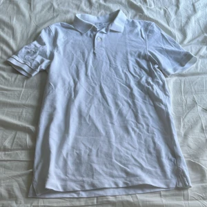 Vit kortärmad pikétröja - Säljer en klassisk vit pikétröja med korta ärmar och krage. Perfekt för en stilren och avslappnad look. Tröjan har knappar framtill och är i ett mjukt material.