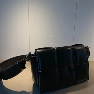 Svart quiltad axelväska - Snygg svart axelväska i quiltad design med bred axelrem. Väskan har ett stilrent utseende och är tillverkad i skinnliknande material. Perfekt för att bära dina dagliga nödvändigheter med stil.