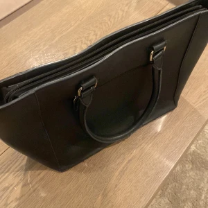 Svart axelväska med dragkedja - Snygg och praktisk svart axelväska i syntetmaterial. Väskan har en dragkedja upptill och flera fack inuti för enkel organisering. Perfekt för vardagsbruk med stilrena handtag och diskreta detaljer.