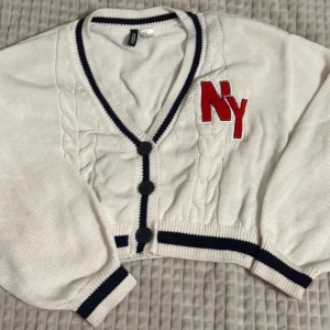 Vit kofta med NY-brodyr - Snygg vit kofta med svart kant och stora knappar. Den har en röd 'NY'-brodyr på bröstet och kabelstickade detaljer. Perfekt för en casual look. Om ni vill se den på, skriv gärna!