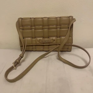 Beige axelväska från Michael Kors - Snygg beige axelväska från Michael Kors med ett flätat mönster. Väskan har en justerbar axelrem och en elegant logotypdetalj framtill. Perfekt för att ge din outfit en stilren touch.