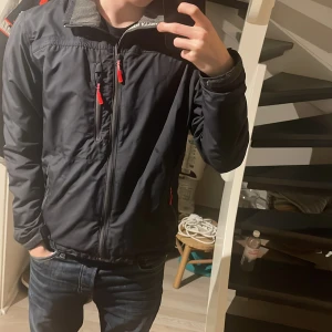 Helly Hansen jacka - Säljer en svart vindjacka från Helly Hansen med röda dragkedjor. Jackan har en hög krage och långa ärmar, perfekt för blåsiga dagar. Den är stilren och funktionell med flera praktiska fickor.xs men passar S