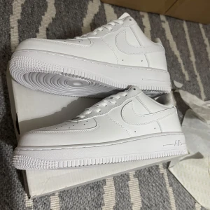 Nike Air Force 1 - Säljer ett par klassiska vita Nike Air Force 1 sneakers. Dessa skor har en stilren design med snörning och är tillverkade i syntetmaterial. Perfekta för en avslappnad och trendig look.