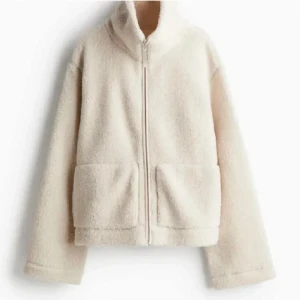 Beige teddyjacka med dragkedja - Mysig beige teddyjacka med dragkedja och hög krage. Jackan har två stora fickor framtill och långa ärmar. Perfekt för kyliga dagar.