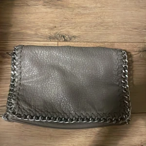 Grå axelväska från Tiamo - Snygg grå axelväska från Tiamo med kedjedetaljer runt kanterna och axelremmen. Säljer på grund av att den inte kommer till användning längre. Den är dock lite sliten på baksidan och dragkedjan på baksidan är sönder. Men inget man tänker på mycket🩷