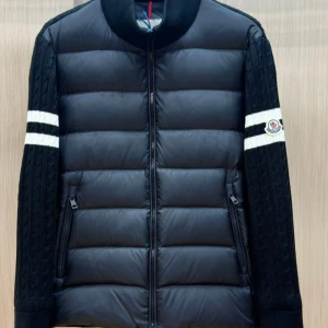 Svart pufferjacka från Moncler - Snygg svart pufferjacka från Moncler med stickade ärmar och vita ränder. Jackan har dragkedja och en broderad logga på ärmen. Perfekt för kyliga dagar.