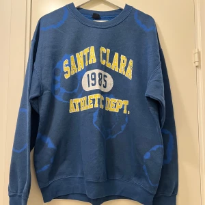 Sweatshirt från Urban outfitters - Blå sweatshirt med texten 'Santa Clara 1985 Athletic Dept.' i gult och vitt på framsidan. Tröjan har ett unikt mönster med ljusare blå cirklar och långa ärmar. Bra skick. Köpt på Urban outfitters men säljs inte längre.