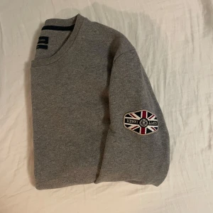Henri Lloyd Tröja - Säljer en stilren grå tröja från Henri Lloyd med ett broderat märke på ärmen. Storlek L. I mycket bra skick bara använd 1 eller 2 gånger