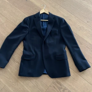 Svart kavaj från Oscar Jacobson - Elegant svart kavaj från Oscar Jacobson med klassisk design. Kavajen har en stilren passform och är försedd med knappar framtill. Fodret är i en mörk nyans med märkets logotyp diskret invävd. Perfekt för formella tillfällen.  