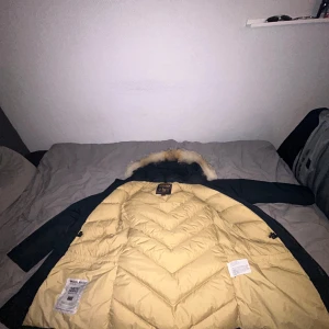 Mörkblå Arctic Parka från Woolrich - Säljer en stilren mörkblå Arctic Parka från Woolrich med avtagbar päls på huvan. Jackan är dunfylld och har knappar framtill. Perfekt för kalla dagar i stort sätt helt ny 