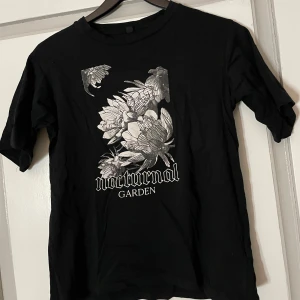 Svart t-shirt med tryck 'Nocturnal Garden' - Snygg svart t-shirt med ett coolt tryck av blommor och texten 'Nocturnal Garden' i vitt. Perfekt för en stilren och mystisk look. Kortärmad och i bekvämt material.