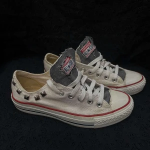 Vita sneakers med nitar från Converse - Unika vita Converse sneakers med nitar på sidan och klassisk snörning. Skorna har en röd rand längs sulan och en grå denim detalj vid plösen. 
