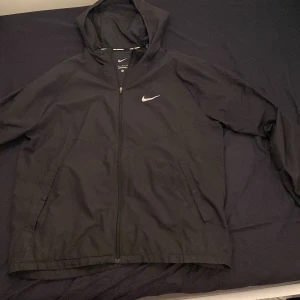 Svart vindjacka från Nike - Säljer en svart vindjacka från Nike med huva och dragkedja. Jackan har långa ärmar och två fickor framtill. Perfekt för löpning och andra utomhusaktiviteter.