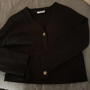 Svart kofta från NA-KD - Snygg svart kofta från NA-KD med v-ringning och dekorativa knappar framtill. Koftan har långa ärmar och ribbade detaljer vid ärmslut och nederkant. Perfekt för en stilren look.
