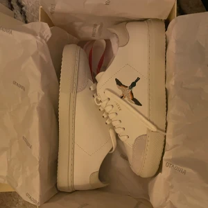 Vita sneakers från Axel Arigato - Original pris:2500kr. Snygga vita sneakers från Axel Arigato med en broderad fågel på sidan. Skorna har en klassisk design med snörning och en bekväm sula. Perfekta för en stilren look. (Säljer då dom va för små)