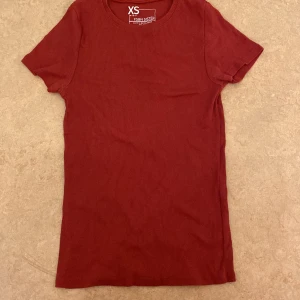 Röd ribbad t-shirt från Gina Tricot - Snygg röd ribbad t-shirt från New Yorker i storlek XS. Perfekt för en casual look med korta ärmar och rund halsringning.