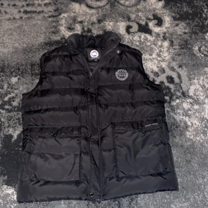 Svart dunväst från Canada Goose - Säljer en svart dunväst från Canada Goose med dragkedja och knappar. Västen har två stora fickor framtill och en broderad logotyp på bröstet. Perfekt för kyliga dagar.  Pris kan diskuteras!    Jag är 183 cm lång och väger 70 den passar perfekt på mig.