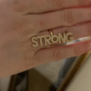 Guldfärgad Ioaku ring ”STRONG” - Trendig guldfärgad ring med texten STRONG i glittriga bokstäver från Ioaku! Passar utmärkt som en unik accessoar till din outfit. Den är i gott skick och är justerbar!