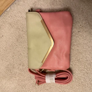 Rosa och beige axelväska med gulddetaljer - Snygg axelväska i rosa och beige med gulddetaljer. Väskan har flera fack med dragkedja och en justerbar axelrem.